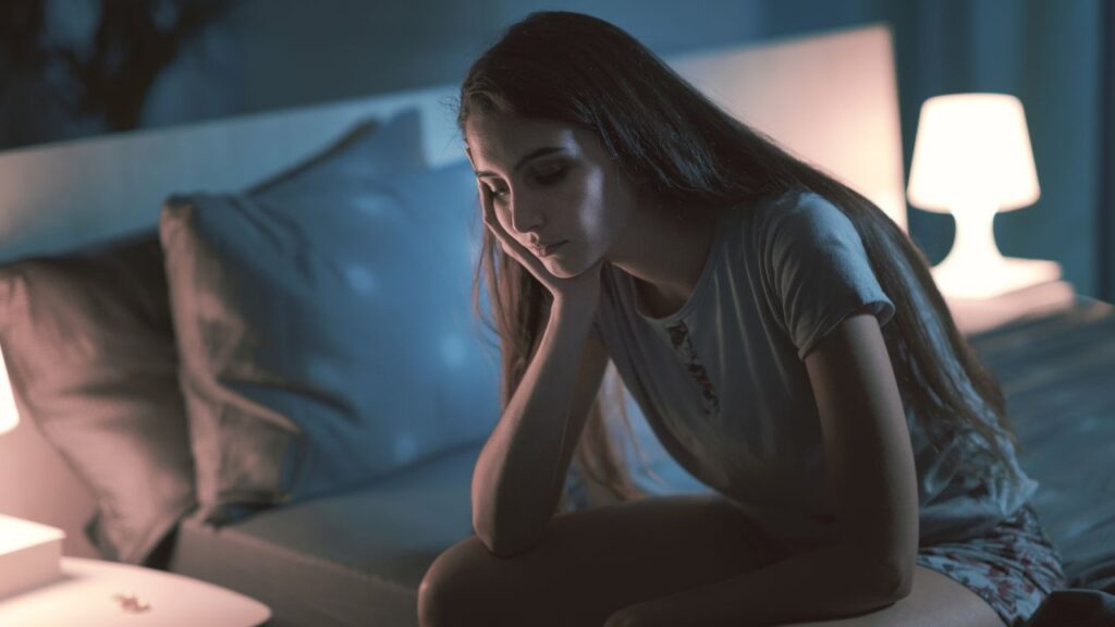 A woman sitting on the edge of her bed in a dimly lit room, appearing tired and restless to illustrate the need for a sleep disorders treatment chiropractor at Spine and Joint Center in Fort Lauderdale.