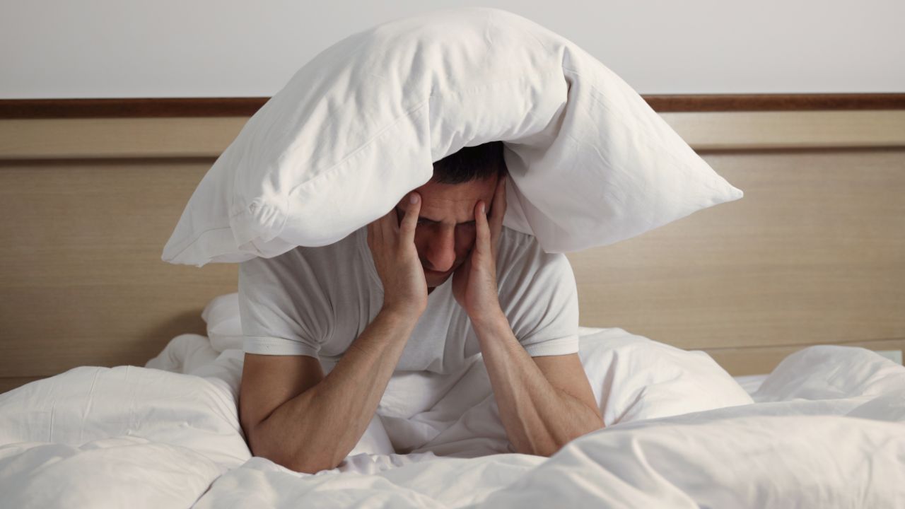 A man sitting in bed with a pillow over his head and his hands on his face to show the frustration of insomnia and the need for a sleep disorders treatment chiropractor at Spine and Joint Center in Fort Lauderdale.