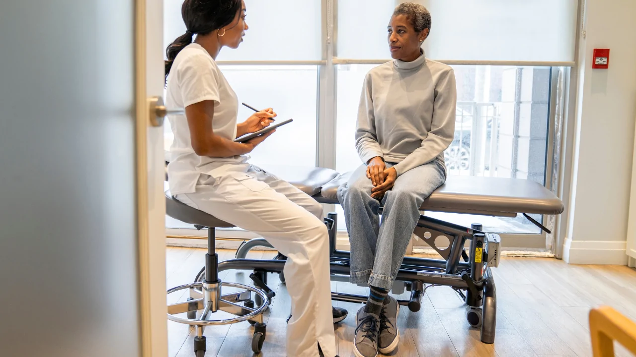 A healthcare provider in white scrubs taking notes during a consultation with a patient in a clinical setting to develop chiropractic exercise programs at Spine and Joint Center in Fort Lauderdale.