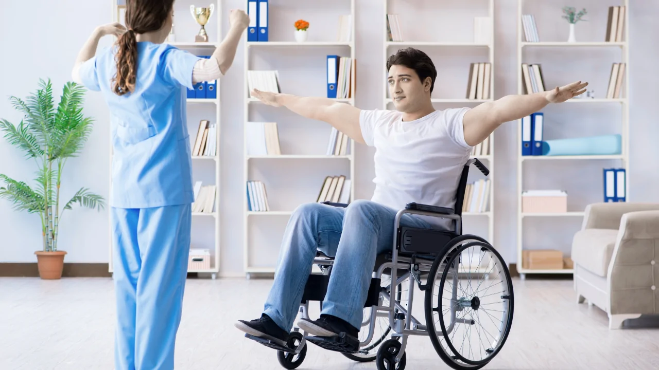 A clinician in blue scrubs guiding a male patient in a wheelchair through bilateral arm extensions to demonstrate inclusive strategies used by an injury prevention chiropractor in Fort Lauderdale.