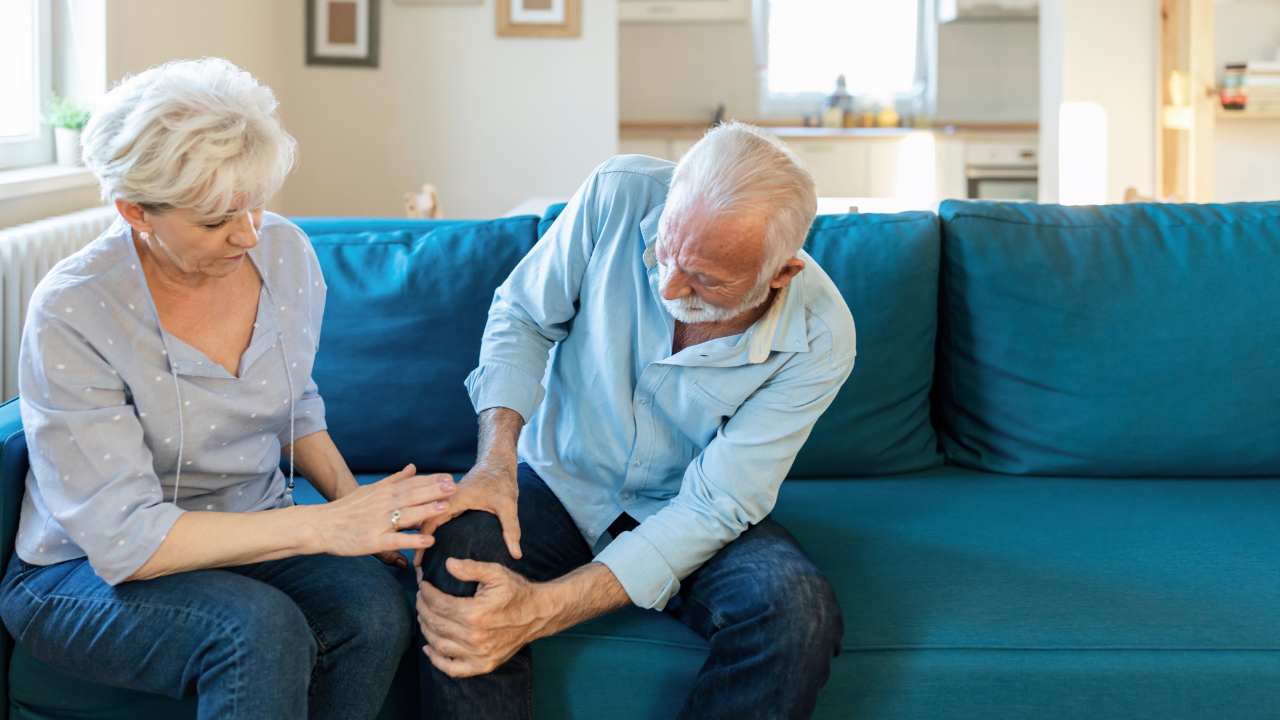 An older man in a light blue shirt sitting on a teal couch and holding his pained knee while a healthcare provider places a comforting hand on his shoulder to illustrate the need for an arthritis pain relief chiropractor at Spine and Joint Center in Fort Lauderdale.