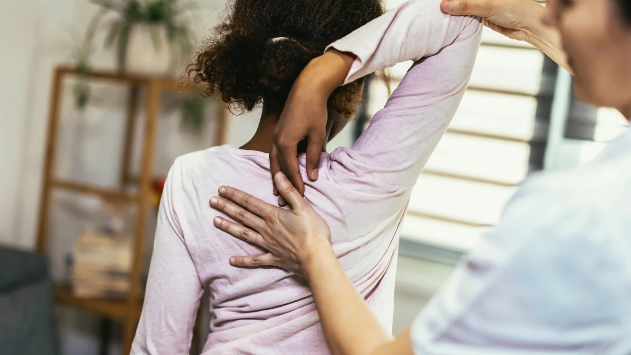 A chiropractor in a gray medical tunic performing a gentle spinal adjustment on the upper back of a young girl to provide pediatric bedwetting treatment at Spine and Joint Center in Fort Lauderdale.
