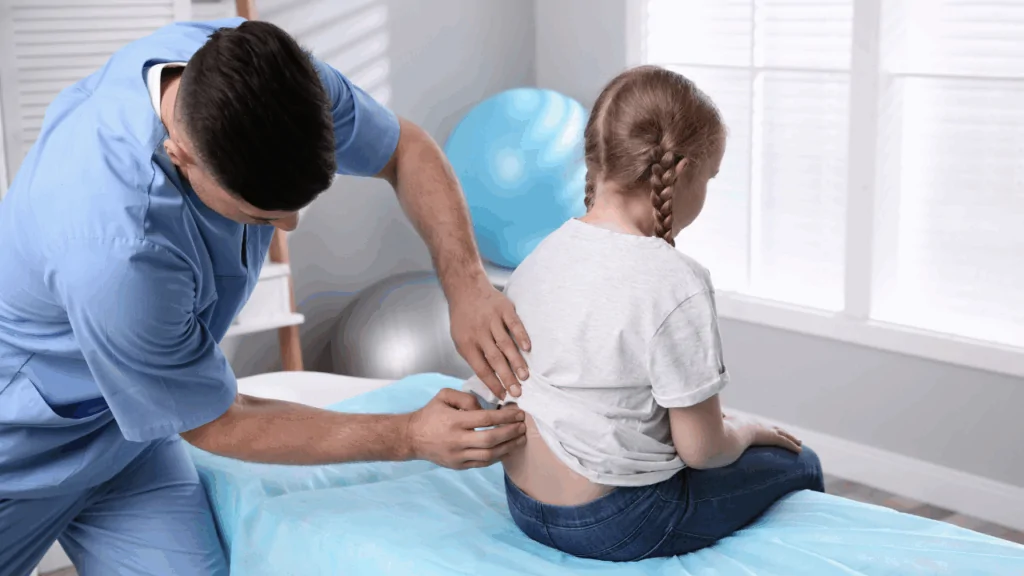 A chiropractor in a blue medical tunic performing a focused spinal palpation on a young girl sitting on a treatment table to represent bedwetting treatment at Spine and Joint Center in Fort Lauderdale.