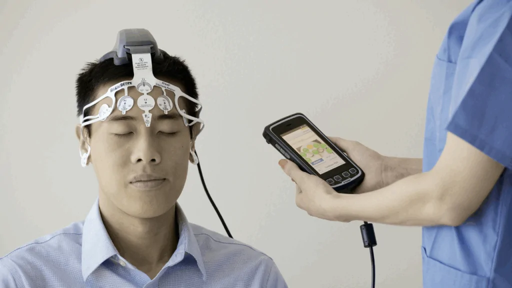 A male patient wearing a specialized head-mounted diagnostic sensor while a clinician monitors data on a handheld device for concussion treatment at Spine and Joint Center in Fort Lauderdale.