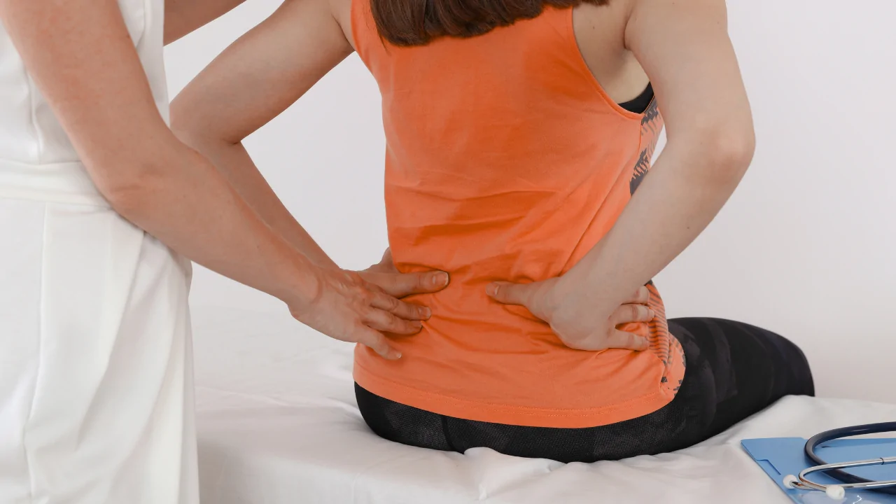 A woman in an orange tank top sitting on a bed and holding her lower back and hip to represent the symptoms treated with hip pain chiropractic care at Spine and Joint Center in Fort Lauderdale.