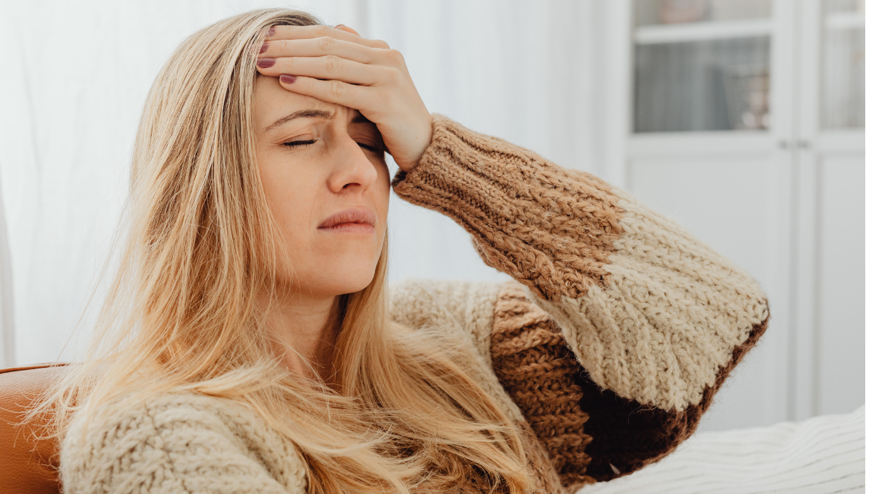 A blonde woman in a tan sweater closing her eyes and holding her forehead in pain to illustrate the need for a migraine and headache chiropractor at Spine and Joint Center in Fort Lauderdale.