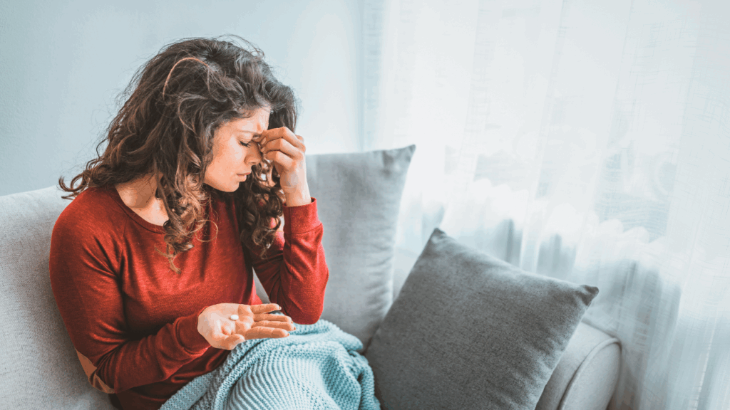 A woman in a red sweater sitting on a couch and holding her head in pain while looking at a white pill in her hand to represent the need for a migraine and headache chiropractor at Spine and Joint Center in Fort Lauderdale.