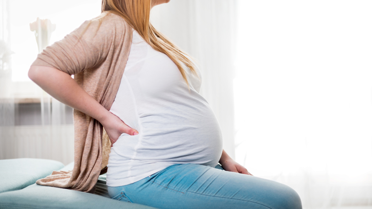 A pregnant woman in a white shirt and tan cardigan sitting and holding her lower back in pain to illustrate the need for pelvic pain during pregnancy treatment at Spine and Joint Center in Fort Lauderdale.