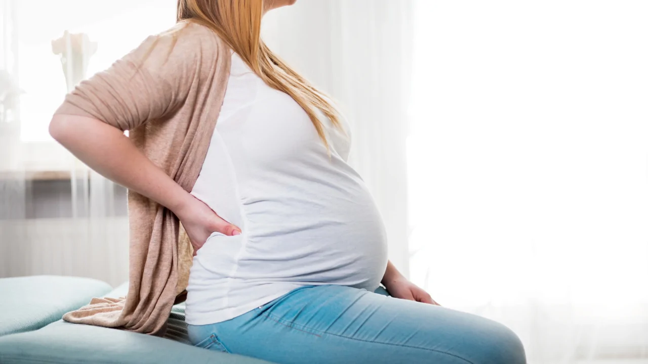 A pregnant woman in a white shirt and tan cardigan sitting and holding her lower back in pain to illustrate the need for pelvic pain during pregnancy treatment at Spine and Joint Center in Fort Lauderdale.