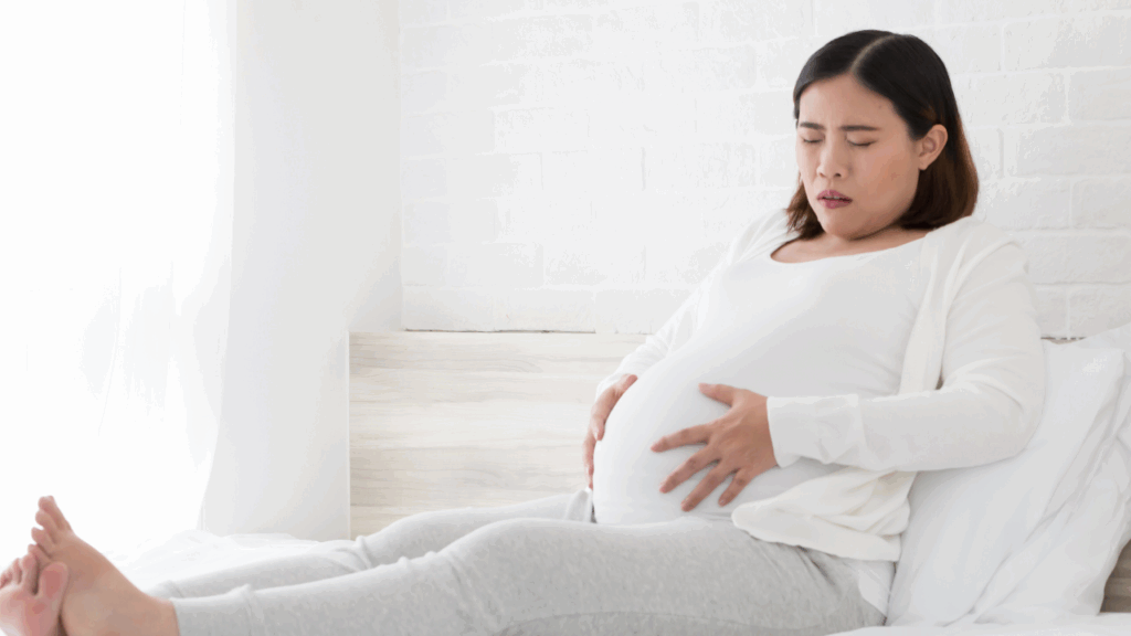 A pregnant woman in a white long-sleeve shirt sitting on a bed and holding her stomach in pain to illustrate the need for pelvic pain during pregnancy treatment at Spine and Joint Center in Fort Lauderdale.