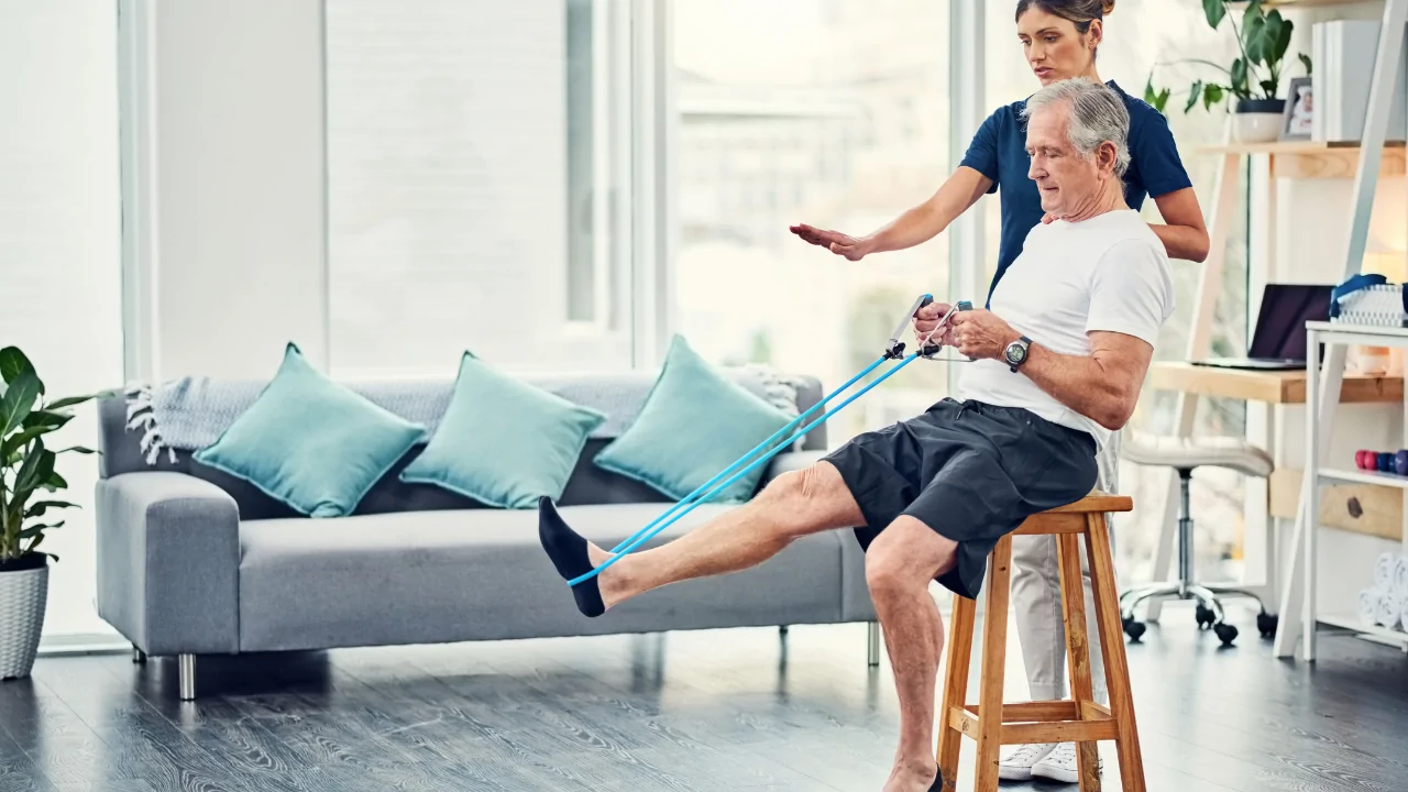 An elderly man sitting on a wooden stool and using a blue resistance band to perform leg extensions under the guidance of a clinician for fall prevention at Spine and Joint Center in Fort Lauderdale.