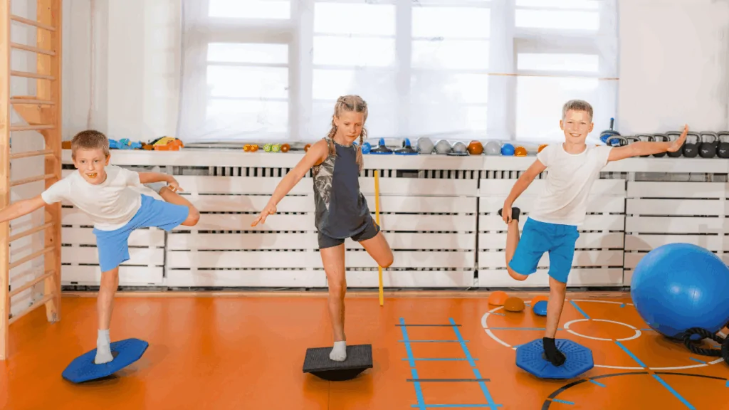 Three young children practicing balance on blue and black wobble boards in a gym setting to demonstrate fall prevention chiropractic techniques at Spine and Joint Center in Fort Lauderdale.