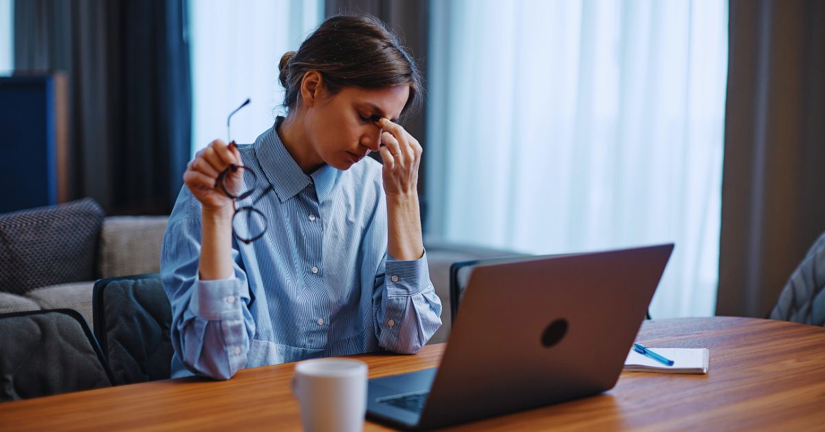 A woman sitting at a desk and rubbing her temples in exhaustion while holding her glasses, highlighting chronic fatigue management services at Spine and Joint Center in Fort Lauderdale.