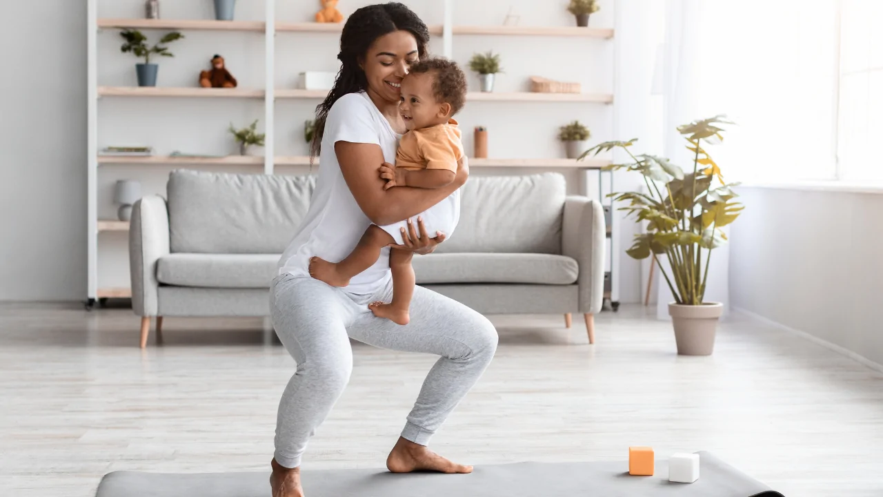 A mother in a white t-shirt and grey leggings performing a guided squat while holding her baby to facilitate postpartum chiropractic recovery at Spine and Joint Center in Fort Lauderdale.