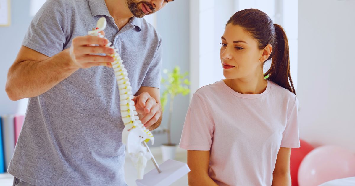 A chiropractor holding a white anatomical spinal model to explain the structural curvature of the spine to a female patient during a session for scoliosis care at Spine and Joint Center in Fort Lauderdale.