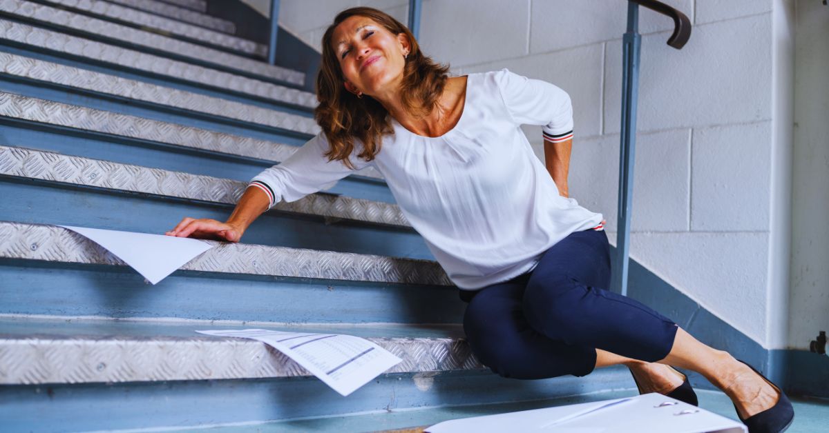 A woman sitting on metal stairs and clutching her lower back in pain after a tumble to show the need for slip and fall injury care at Spine and Joint Center in Fort Lauderdale.
