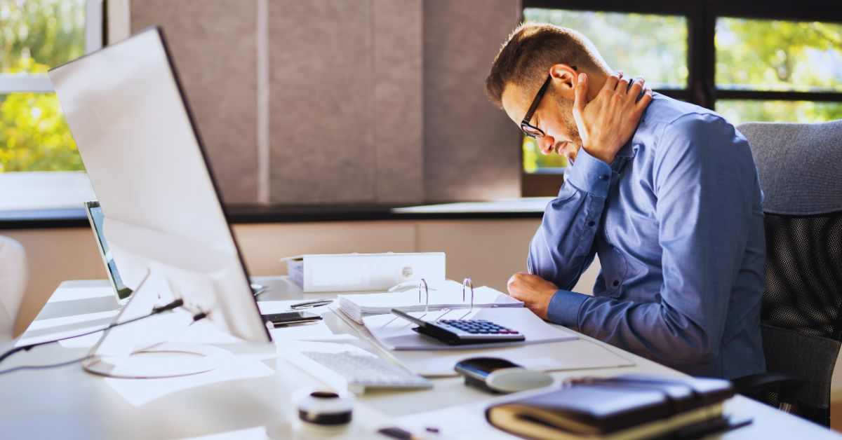 A man sitting at an office desk holding his neck in pain due to poor posture, highlighting the need for professional workplace ergonomics and pain relief in Fort Lauderdale.